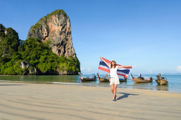 Una donna con la bandiera thailandese cammina su una spiaggia di Krabi, con le barche a coda lunga e un torrione di roccia alle spalle